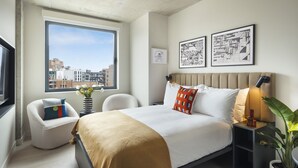 A modern bedroom with a large window, a bed with a beige blanket, a chair with a striped pillow, and two framed pictures on the wall. - Penny Williamsburg (Brooklyn)