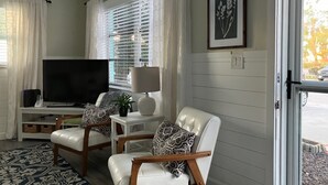 Un salón con un televisor de pantalla plana grande, dos sillones de cuero blanco, una mesita con una lámpara e impresión botánica enmarcada en la pared.