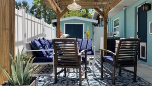 Un patio al aire libre con una pérgola de madera, alfombra estampada en azul y blanco, dos sillas de madera, un sofá blanco con cojines azules y una mesita.