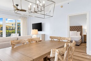 Une chambre d’hôtel avec une table à manger en bois, un canapé, un lit doté d’une tête de lit capitonnée, un téléviseur fixé au mur et une grande fenêtre donnant sur des immeubles.
