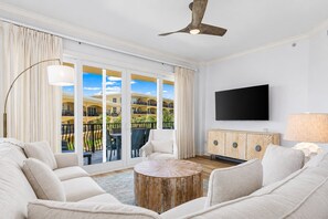 A modern living room with a sofa set, a round coffee table, a television mounted on the wall, and a balcony with a view of buildings and greenery. - Sea View Serenity - 3BR/3BA Adagio Condo - Sleeps 8 - Community Pool View (Santa Rosa Beach)