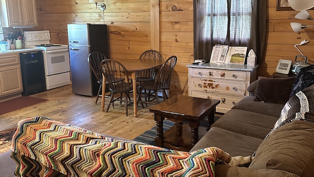 A cozy cabin interior with a wooden ceiling, a dining area, a kitchenette, and a living room with a couch and a colorful blanket. - Lyman Run Cabin - 2 miles from Lyman Run State Park - on ATV trails (Galeton)