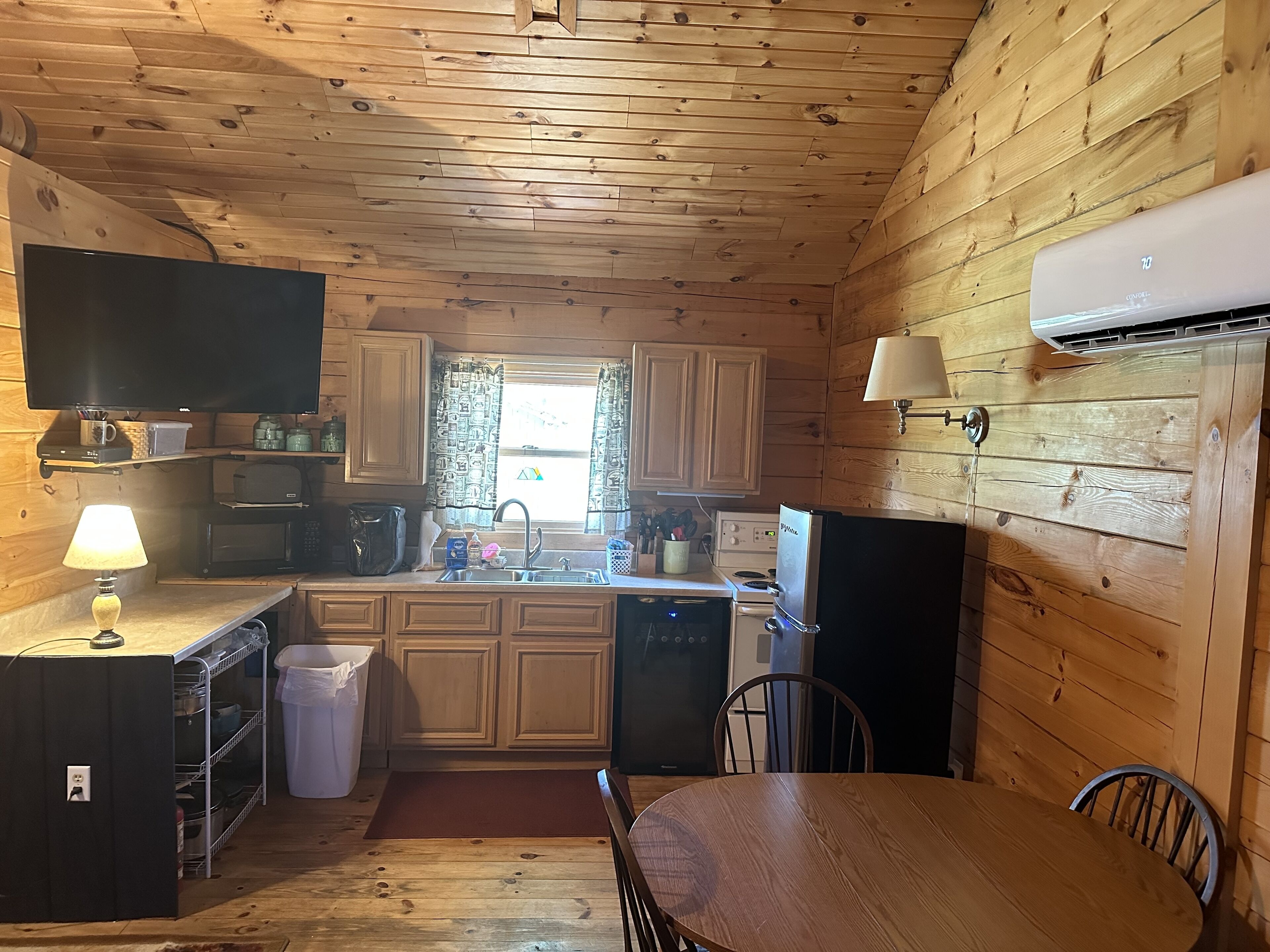 A compact kitchen with wooden walls and ceiling, a microwave, stove, sink, and a dining area with a table and chairs.