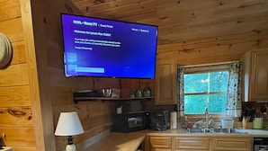 A cabin interior with a TV displaying a welcome message, wooden cabinets, a microwave, a sink, and a window with a view of trees. - Lyman Run Cabin - 2 miles from Lyman Run State Park - on ATV trails (Galeton)