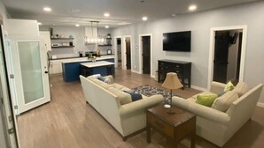 A modern living room with a sofa, coffee table, and a patterned rug. There is a kitchen area with a fridge, microwave, and a TV mounted on the wall.