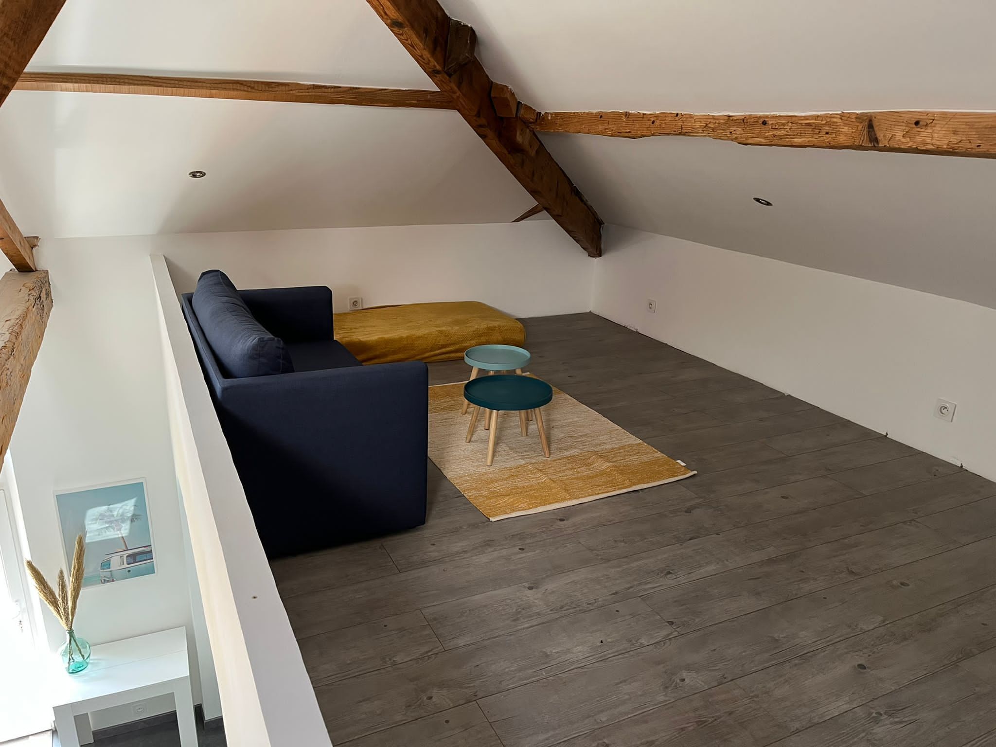 A loft-style room with a dark blue sofa, a mustard-colored ottoman, and two small teal tables.