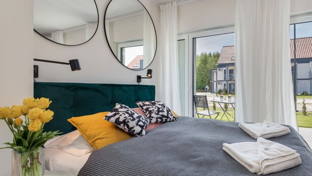 Une chambre à coucher moderne avec un grand lit, une table de chevet ornée d’un vase de fleurs et une vue sur l’extérieur.