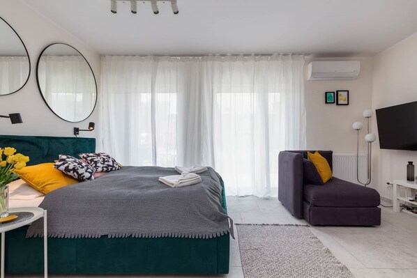 Une chambre moderne avec un grand lit, un canapé, une télévision et une fenêtre ornée de rideaux légers.
