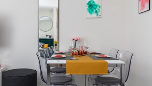 Un espace repas moderne avec une table blanche dressée pour six personnes, des chaises transparentes et un pouf noir.