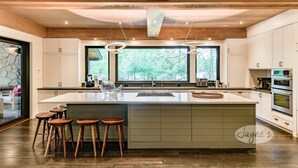 A modern kitchen with a large island, wooden stools, and a view of trees through the windows.