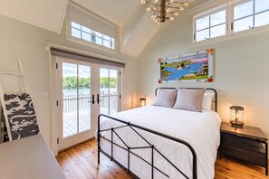 A bedroom with a bed, a painting, a nightstand, a lamp, and a view of a dock and water through a sliding door.