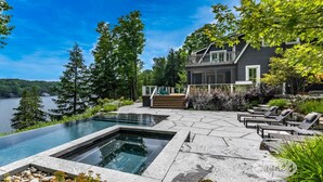 Una casa moderna con una gran piscina exterior, una terraza con sillones de descanso y vistas a un lago.