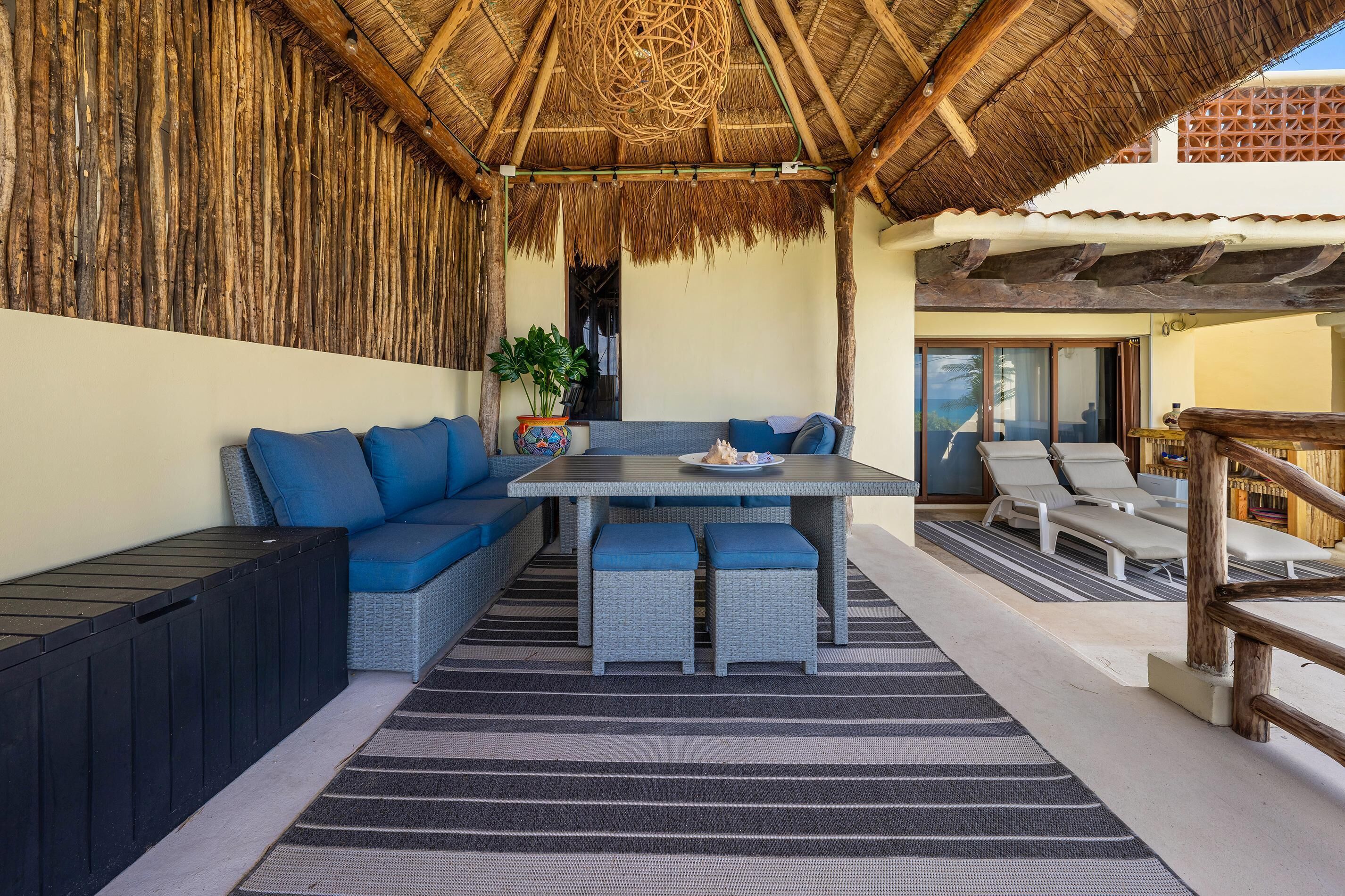 A covered outdoor seating area with a table, cushioned seating, and a thatched roof.