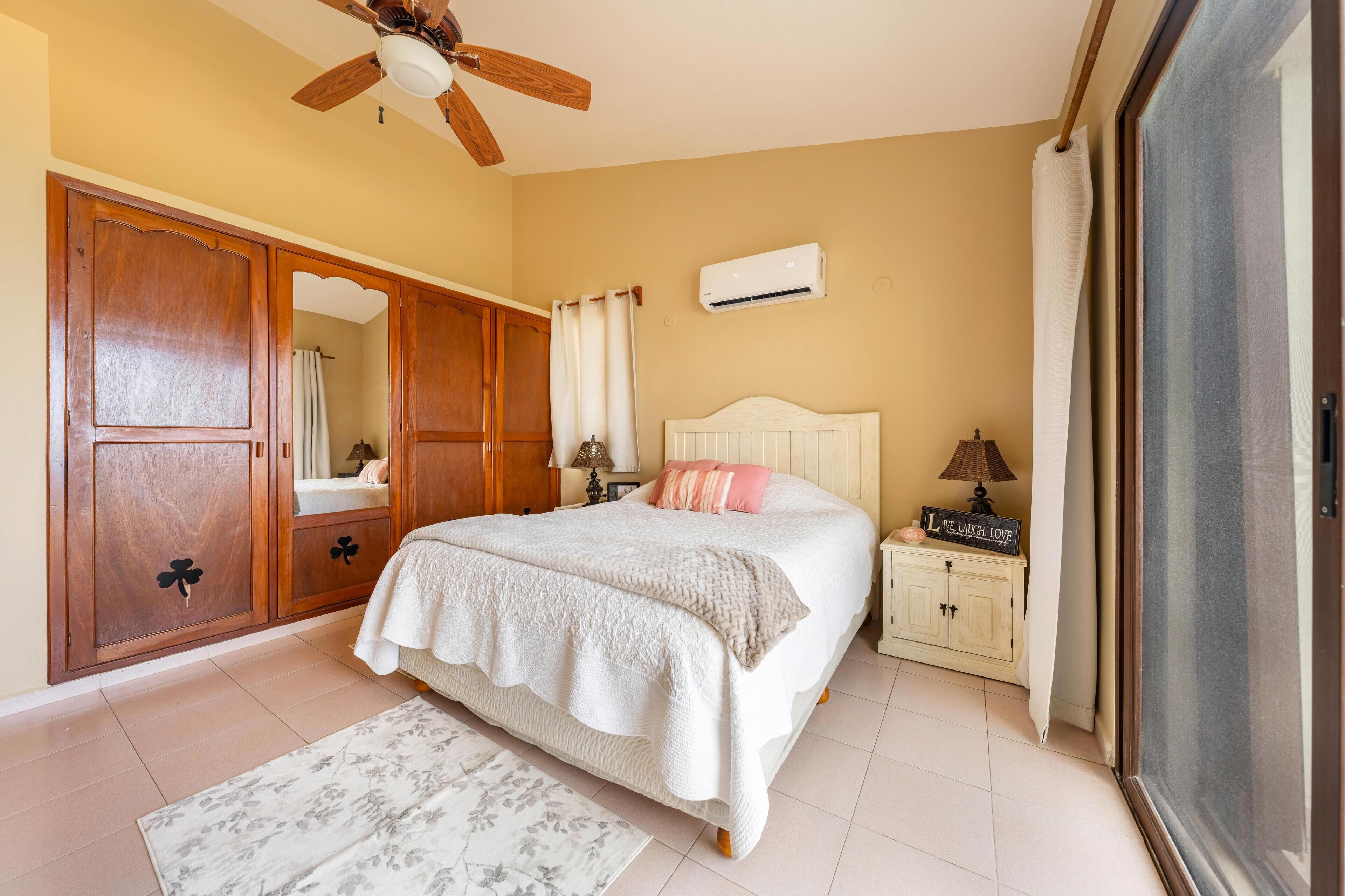 Un dormitorio con cama, mesita de noche, armario, ventilador de techo, aire acondicionado y ventana con cortinas.