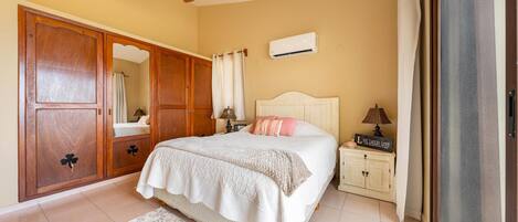Un dormitorio con cama, mesita de noche, armario, ventilador de techo, aire acondicionado y ventana con cortinas.