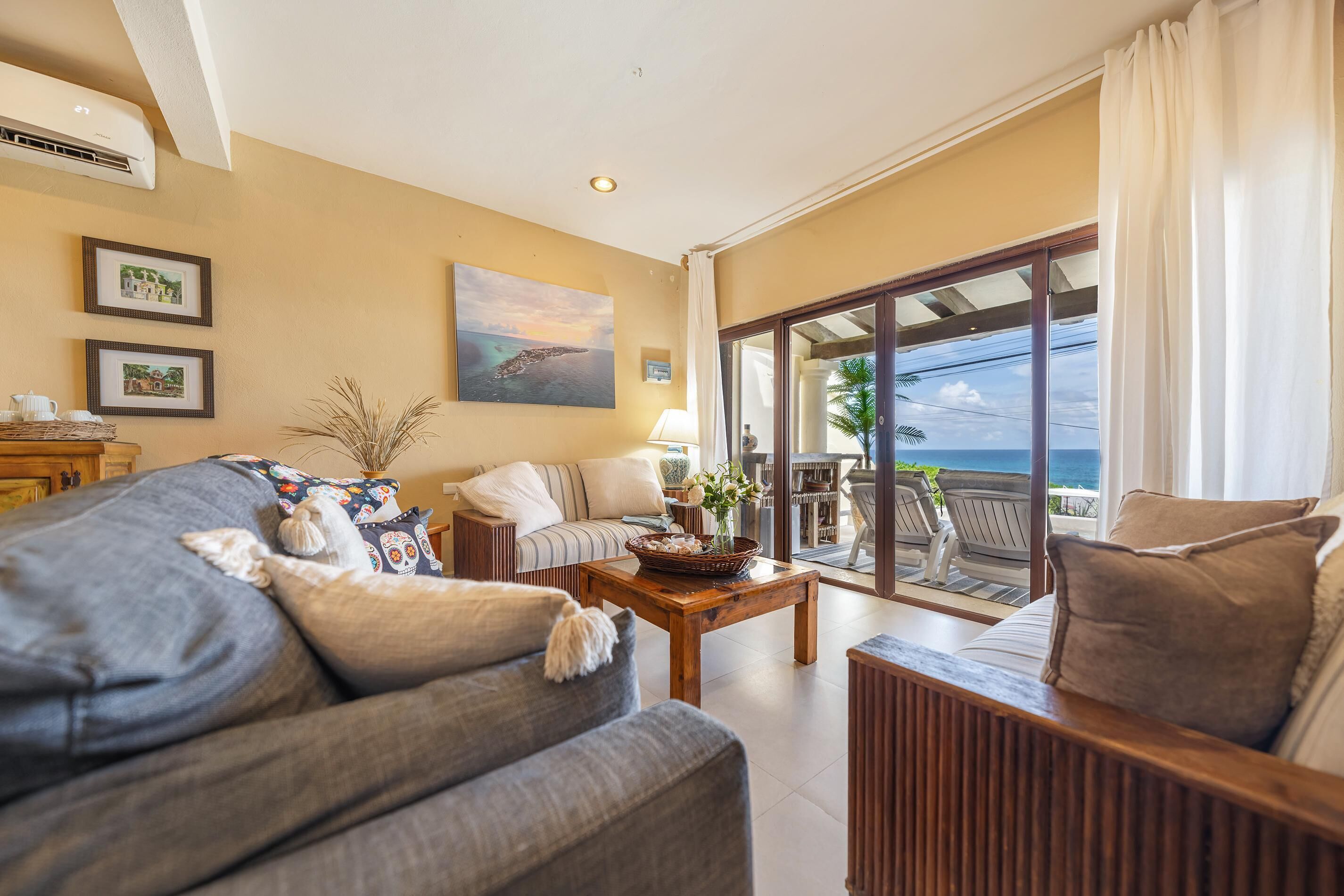 A spacious living room with a sofa, armchair, and a coffee table. There are framed pictures on the wall and a large window with a view of the ocean.