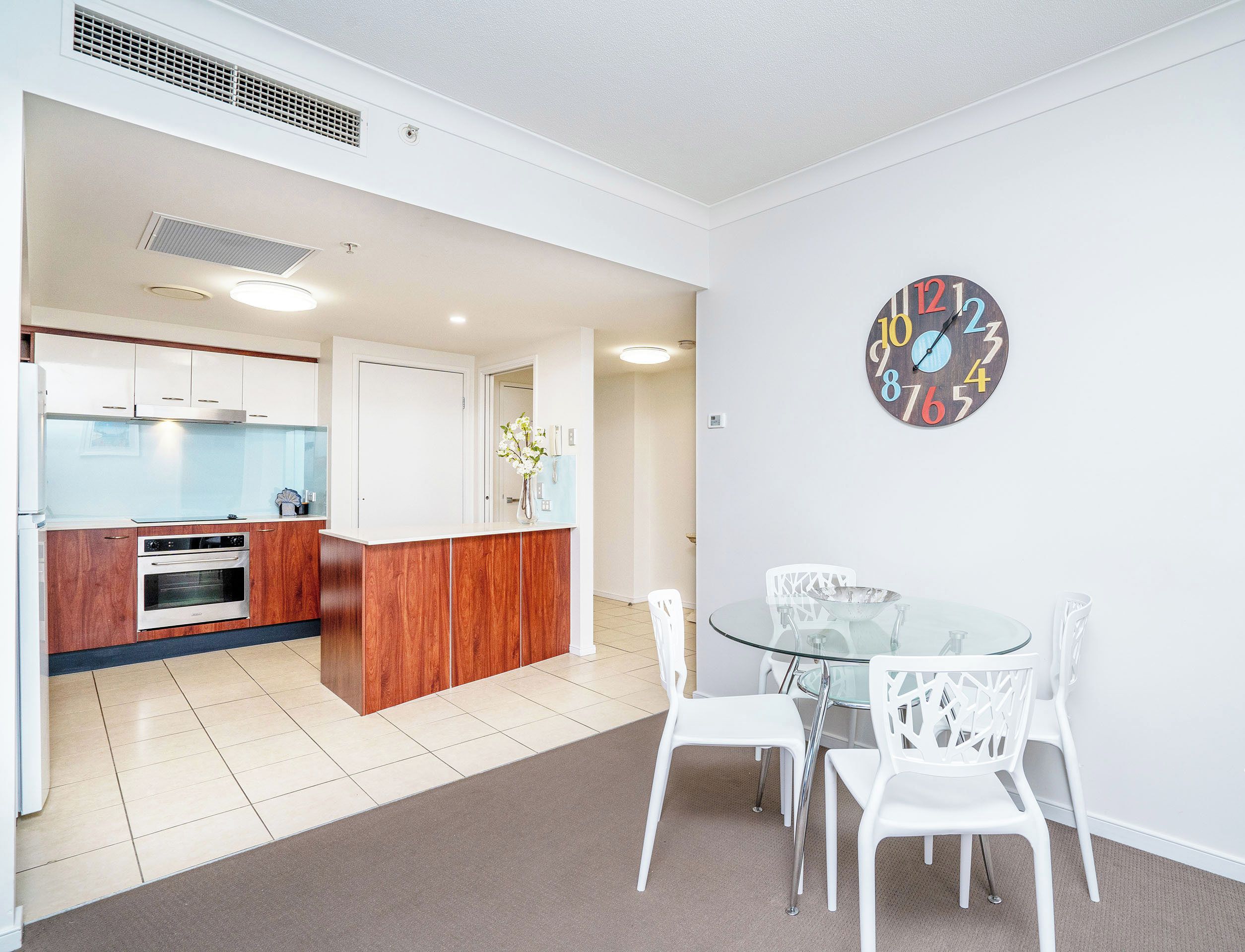 A modern kitchen with white cabinets, a wooden countertop, and a built-in oven. There is a glass dining table with white chairs and a decorative clock on the wall.