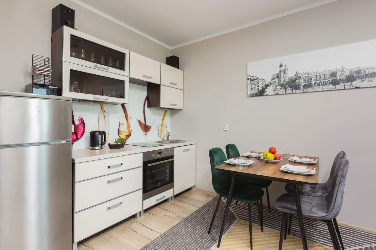 Cocina moderna con armarios blancos, un refrigerador de acero inoxidable y una zona de comedor con mesa de madera y sillas verdes.