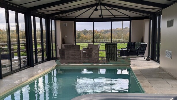 Une piscine extérieure couverte, dotée d’un jacuzzi, entourée de murs de verre et offrant une vue sur la campagne.