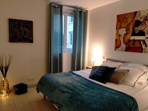 Et soveværelse med seng, natbord, lampe, et maleri og et vindue med gardiner.