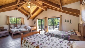A spacious loft bedroom with high ceilings and exposed wooden beams, featuring two beds with patterned quilts, a sofa, and large windows offering a view of the outdoors. - A Resort like Destination & Sleeps 16 Comfortably  (Big Bear Lake)
