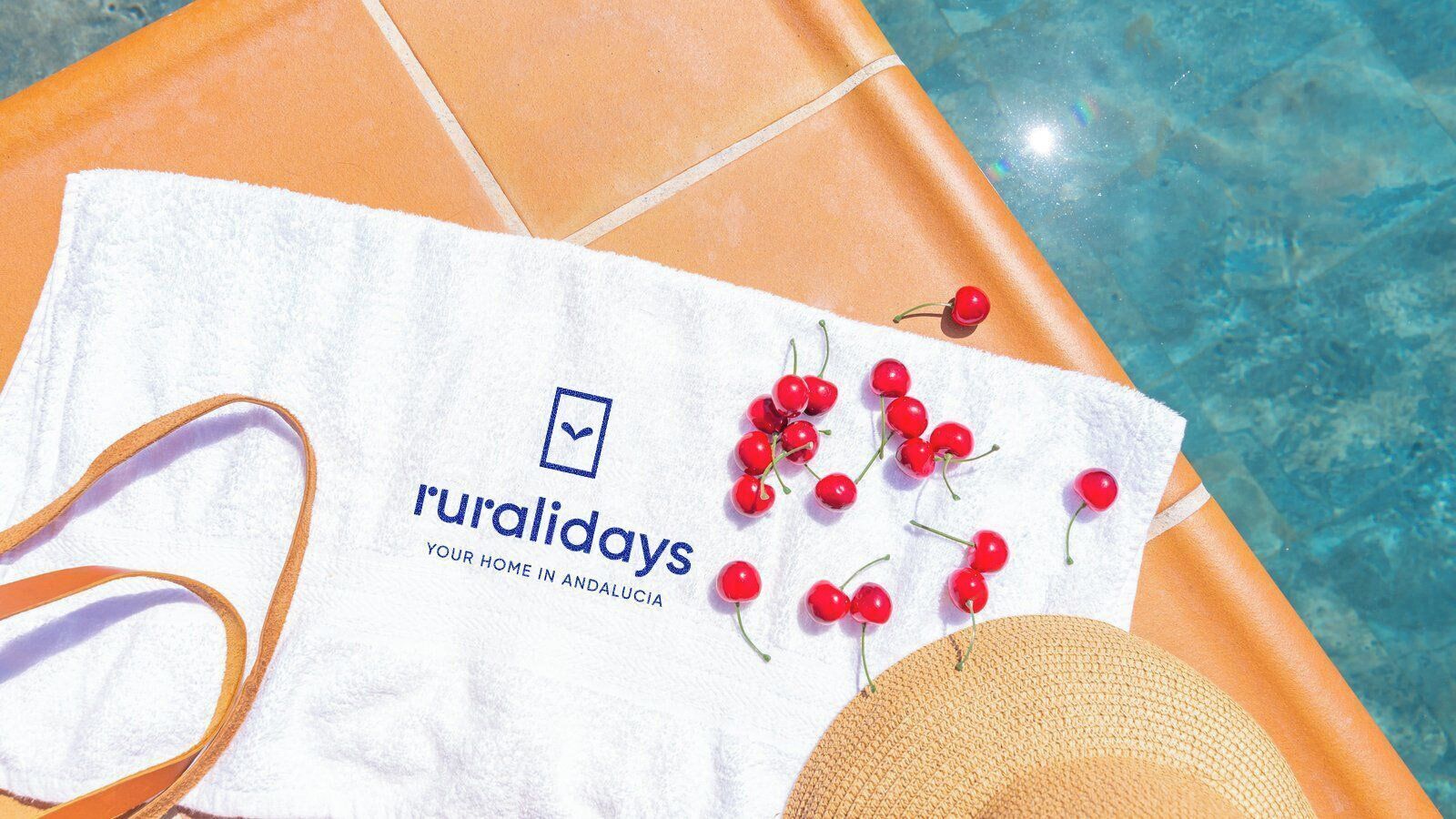 Une serviette ornée du mot « ruralidays », un chapeau de paille et des cerises rouges sur une surface carrelée près d’une piscine.