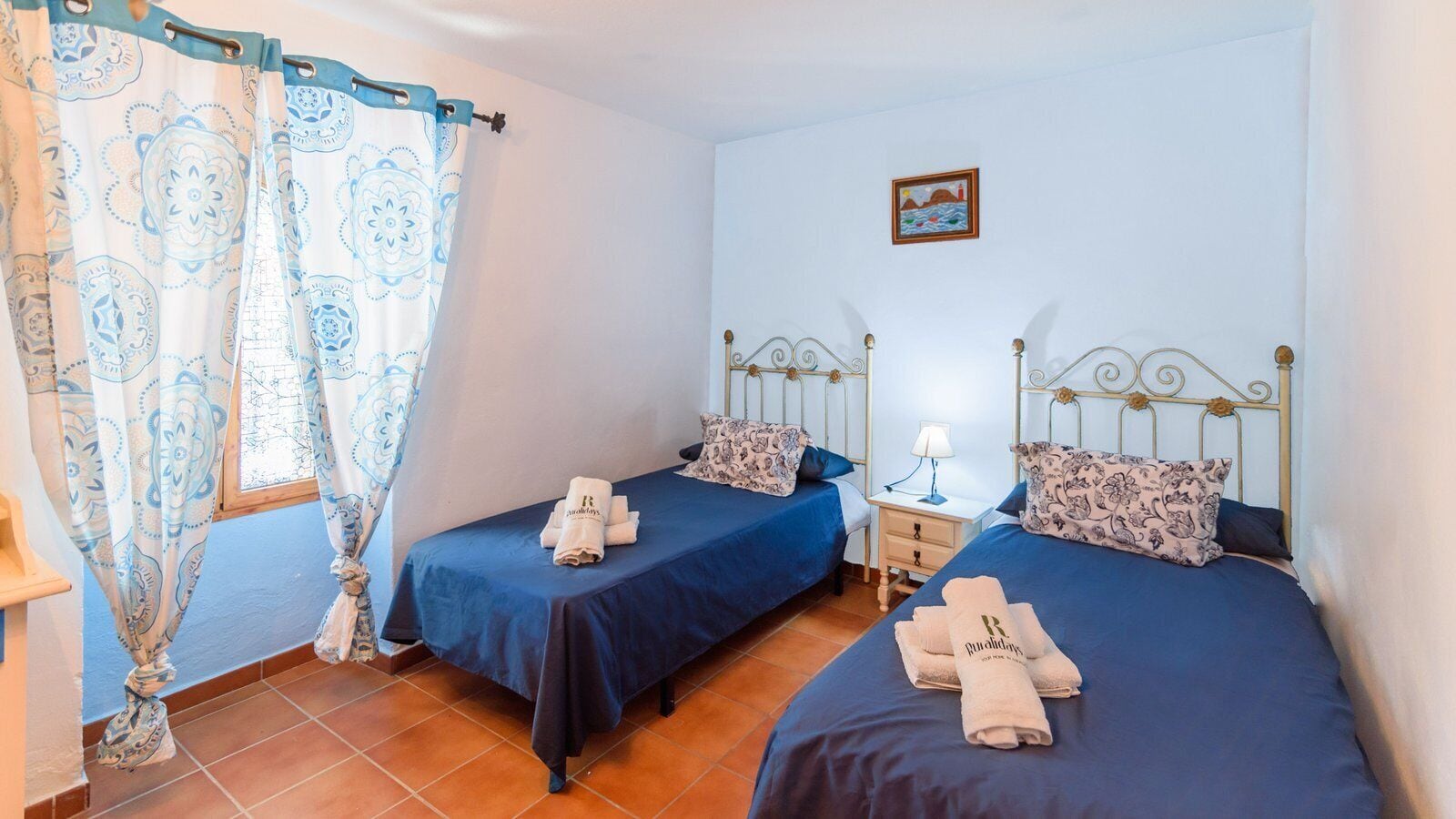 A room with two beds, each with a blue bedspread and matching pillows, a small bedside table with a lamp, and a window with patterned curtains.