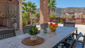 Une terrasse extérieure avec une table dressée pour le repas, entourée de chaises, d’une plante en pot et d’un saladier de fruits.