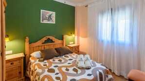 Una cama bien hecha con una colcha estampada, dos almohadas y dos toallas con un logotipo. Se observa un cabecero de madera, dos mesitas de noche con lámparas y un cuadro en la pared.