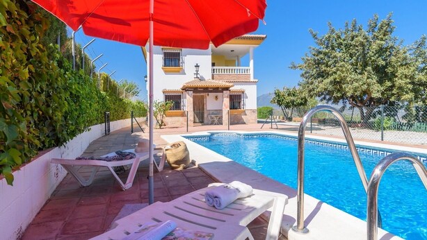 Un área junto a la piscina con sillones de descanso, una sombrilla roja y un edificio al fondo.