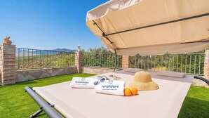 A sunbed with a canopy, a hat, sunglasses, and towels with a logo, set on a green lawn with a mountain view. - Casa Trigueros Alozaina by Ruralidays (Málaga)