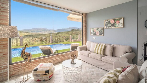 A living room with a large window overlooking a pool and mountains, a beige sofa, and a small round table. - Casa Trigueros Alozaina by Ruralidays (Málaga)