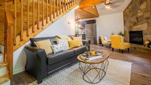 A modern living room with a black leather sofa, a yellow patterned armchair, a wooden staircase, a stone fireplace, and a kitchen in the background. - BEES KNEES A-frame Cabin for 2 * hot tub wifi (Gatlinburg)