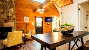 A cozy room with a stone fireplace, wooden walls, a dining table with chairs, and a television displaying the Disney+ logo. - BEES KNEES A-frame Cabin for 2 * hot tub wifi (Gatlinburg)