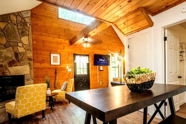 Um quarto acolhedor com lareira de pedra, paredes de madeira, uma mesa de jantar com cadeiras e uma televisão exibindo o logotipo do Disney+.