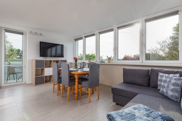 A modern living room with a grey sofa, a dining table with chairs, a TV mounted on the wall, and large windows with a view of greenery.