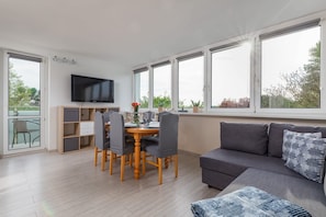 A modern living room with a grey sofa, a dining table with chairs, a TV mounted on the wall, and large windows with a view of greenery.