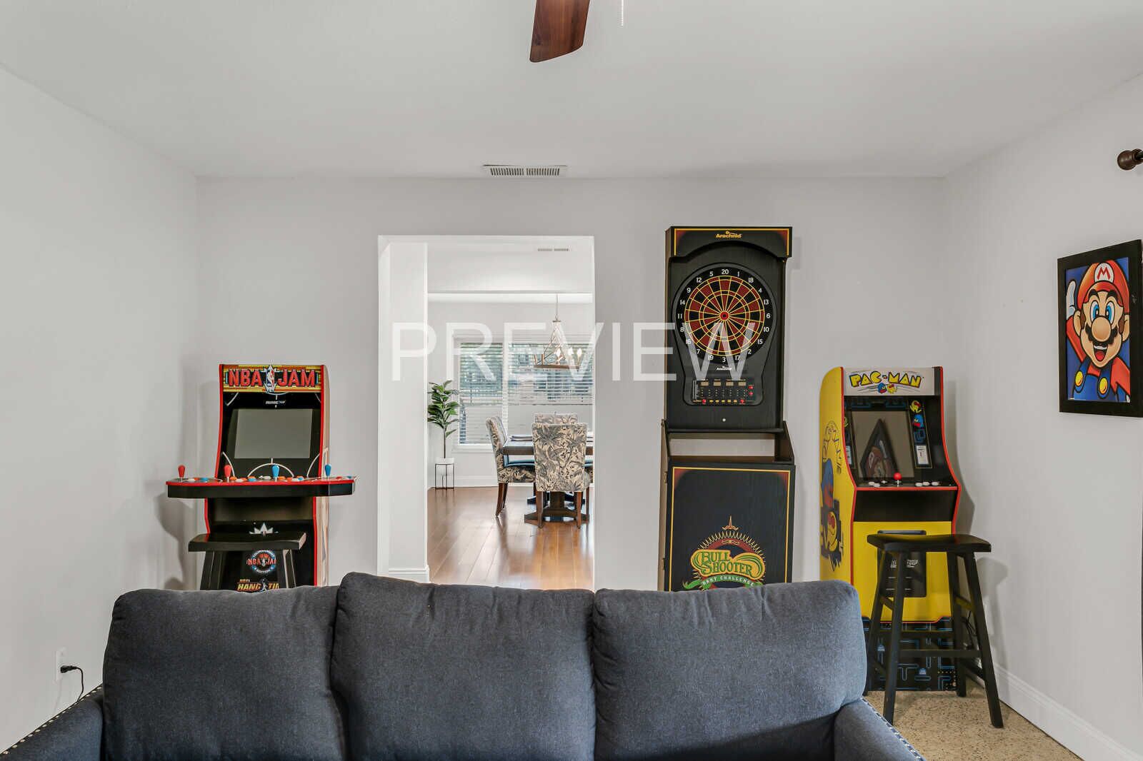 A living room with a grey sofa, an NBA JAM arcade game, a Bull Shooter arcade game, and a Pac-Man arcade game.