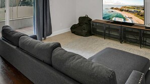 A modern living room with a grey sofa, a TV on a wooden stand, and a large window with curtains. - Spacious Ranch Home w/ Large Yard and Wi-Fi (Tampa)