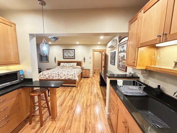A bedroom with a bed, a small sitting area with a stool, a kitchenette with a microwave, and a dining area with a table and chairs.