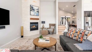 A living room with a fireplace, a flat-screen TV, a round coffee table, and a sofa with patterned cushions.