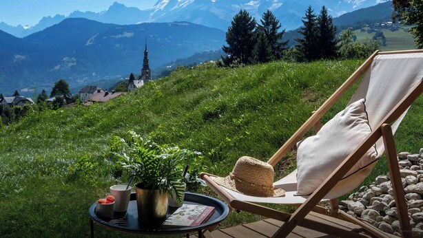 Una silla de descanso con cojín, un sombrero de paja, una planta en maceta y una mesita con tazas y un libro, con vistas a un paisaje montañoso que incluye una iglesia.