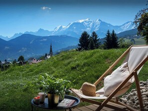 Una cadira de relax amb coixí, un barret de palla, una planta en test i una taula petita amb tasses i un llibre, amb vista a un paisatge montanyós que inclou una església.