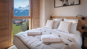 Un dormitorio con una cama grande, ropa de cama blanca, cabecera con lámpara y vista a la montaña a través de una ventana.