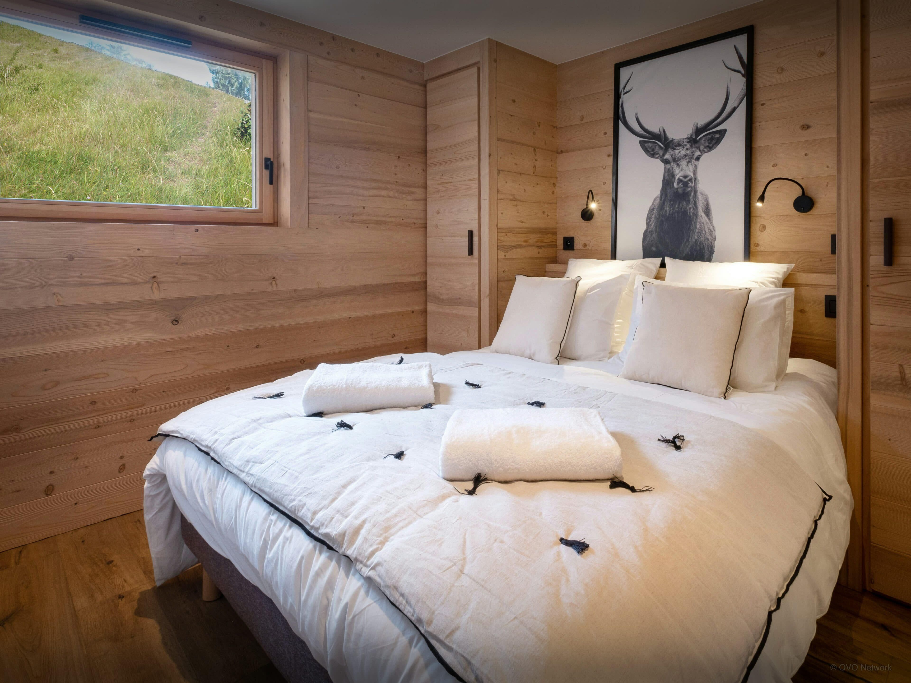 A wooden-paneled bedroom with a large bed, white bedding, a framed deer picture, and a window with a view of greenery.
