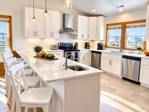 A modern kitchen with white cabinetry, a granite countertop, stainless steel appliances, and a large window offering a view of houses. - Cape Coral 🌊 1-Min Beach | Heated Pool | 🛺 Golf Cart | Elevator | Ocean Views! (Nagshead)