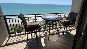 Un balcon avec deux fauteuils en osier et une table à plateau en verre, donnant sur une plage et l’océan.