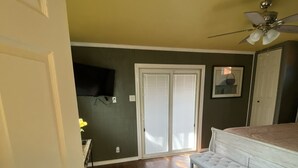 Una habitación con piso de madera, una cama blanca con cabecera, ventilador de techo y puerta corrediza de vidrio que da a un balcón.