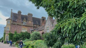 A historic building with a gravel pathway, lush greenery, and a large tree in the foreground. - Six on Castle (Tiverton)