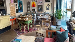 A cozy, well-lit dining area with a large wooden table and chairs, surrounded by various artworks and a menu board. - Six on Castle (Tiverton)